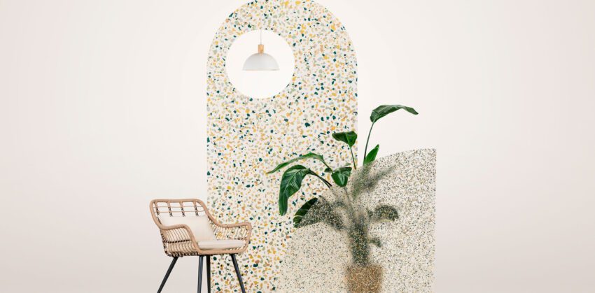 A small vignette on a blank background. An arched panel of translucent resin embedded with colorful fragments is at the back. The panel has a circular cutout that reveals a white hanging light. In front of the panel is a wicker chair and a potted plant with large leaves. There is a smaller resin panel with a dense pattern of small color pieces in the foreground.