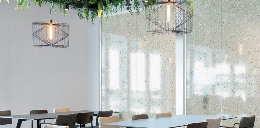 A close up photo of an office break room or dining area. Wire light fixtures hang above tables. The translucent resin inserts in the rear partition wall have a sand-like pattern scattered throughout them.