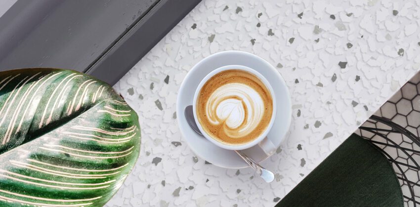 A top down view of a cafe bar. The white and gray bar top has a pattern similar to terrazzo. There is a cup of coffee resting on a saucer on top of the bar, and the leaf from a large indoor plant hangs over the bar as well.